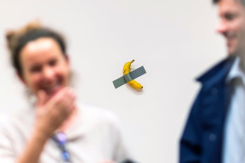 The blurred image of a woman and a man laughing in the foreground with a the focus on a yellow banana taped to a white wall with silver duct tape in the background.