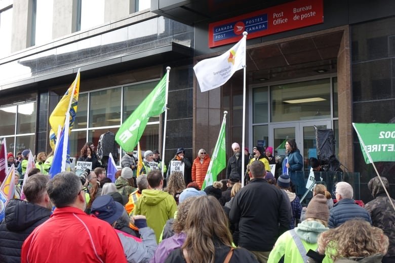 A crowd of people, several of whom are holding flags, listen to a man speaking at a microphone.