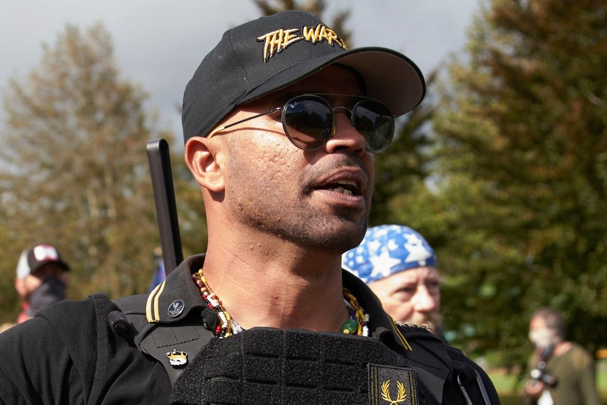 A man wearing a dark baseball cap and sunglasses, sporting some stubble, is shown in an outdoor photo.