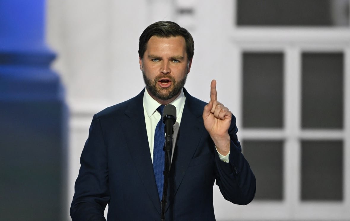 A man in a dark suit points upward while speaking at a microphone.